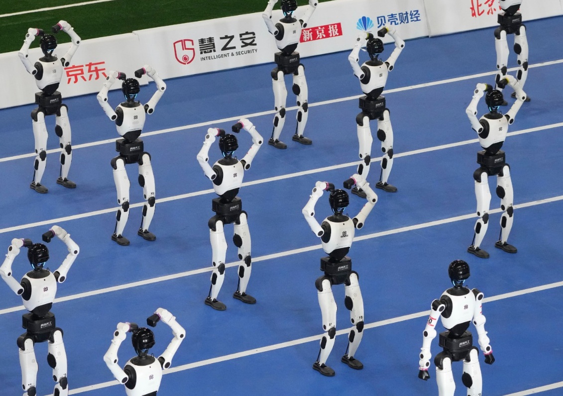 Group of white and black humanoid robots on a blue sports court performing synchronized arm poses.