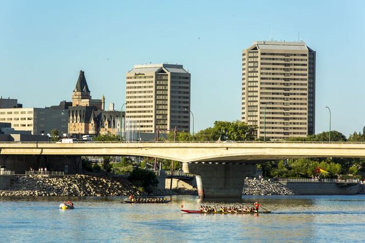 Rowing team paddling a red boat on a river beneath a bridge with a city skyline in the background, including tall office buildings.