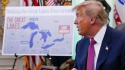 Side view of a man in a navy suit and pink tie, facing a large map labeled The Great Lakes in a formal room.