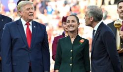 Three people in formal attire stand on a stage; left is a man in a navy suit and red tie, center a smiling woman in a green coat with a flower pin, right a man in a dark suit. A blurred crowd behind them.