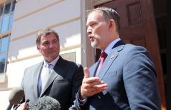 Two men in suits speak to reporters outside a building, microphones in front of them.