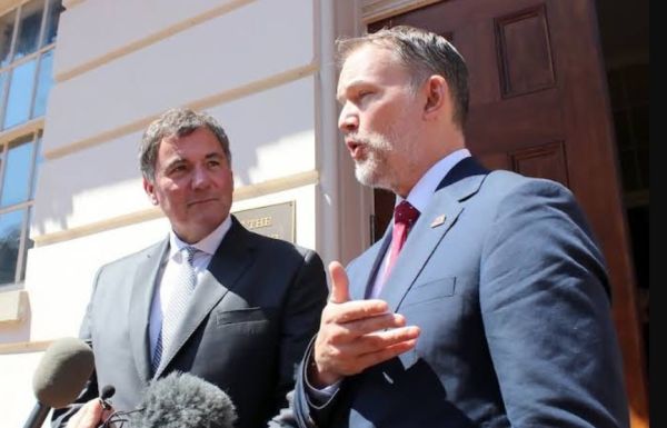 Two men in suits speak to reporters outside a building, microphones in front of them.