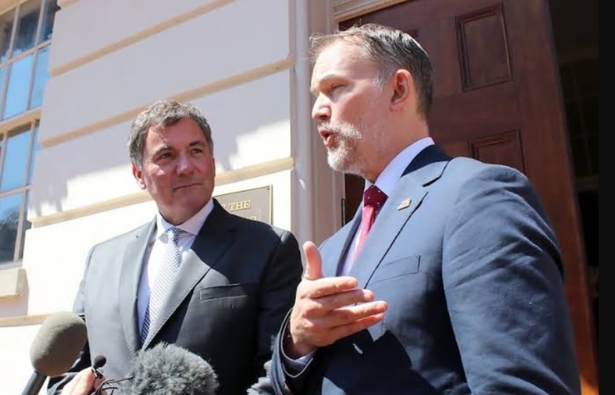 Two men in suits speak to reporters outside a building, microphones in front of them.