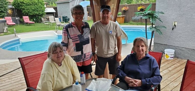 Four seniors posing around a table on a wooden deck beside a backyard pool, smiling, with outdoor chairs and potted plants in the background.