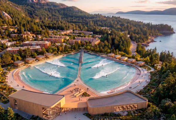Aerial view of a circular wave pool with a central spine bridge at a forested coastal resort, surrounded by buildings and hills.