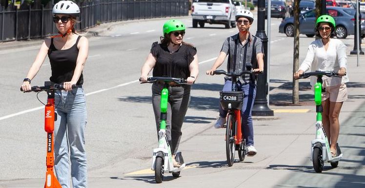 Four people ride electric stand-up scooters on a city street, all wearing helmets and sunglasses.