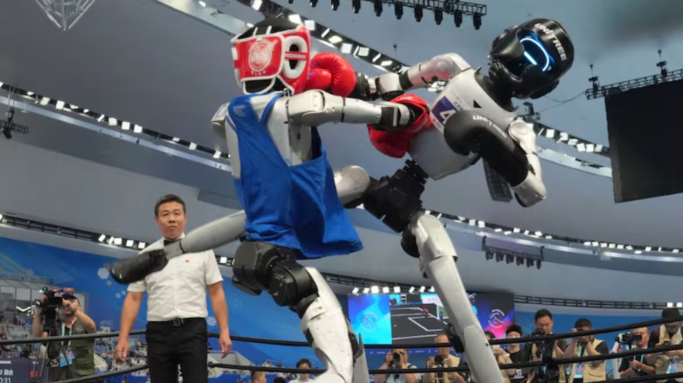 Two humanoid boxing robots clash in a ring as a referee watches and photographers capture the match.