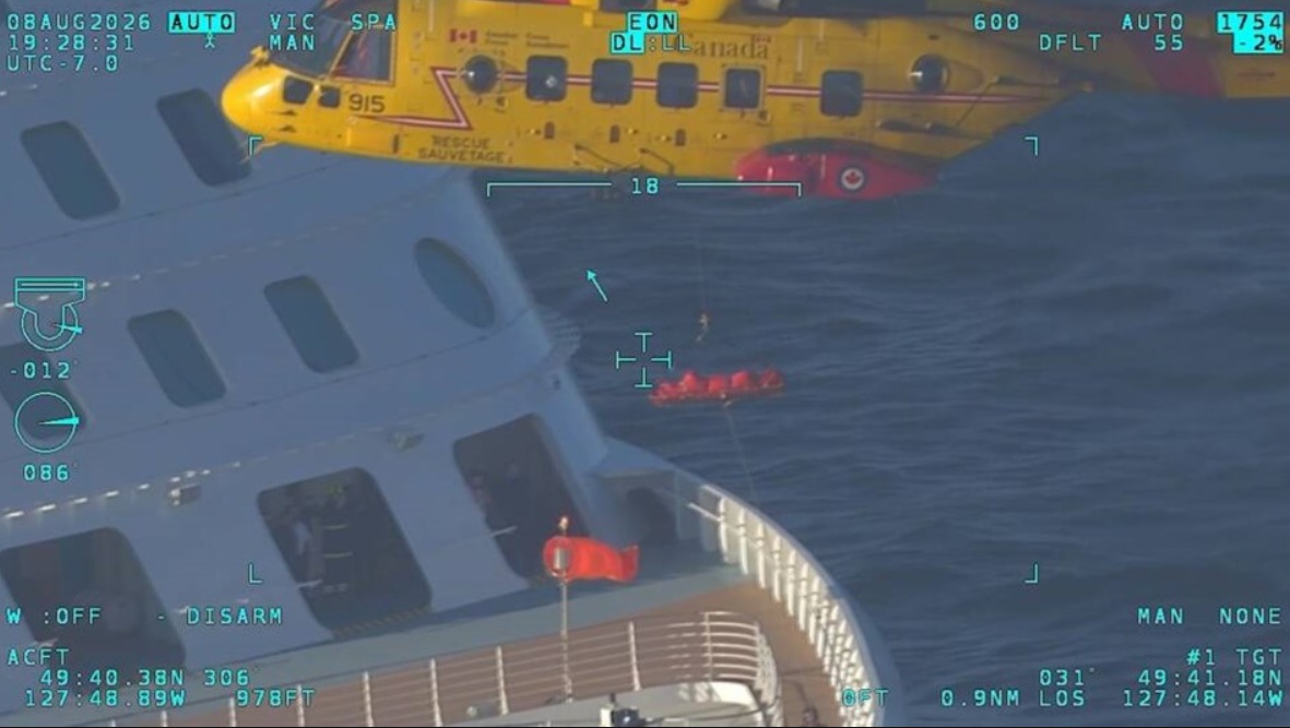 Aerial view from a helicopter HUD showing a large gray ship tilting, with a yellow rescue helicopter above and orange liferafts in the blue water below.