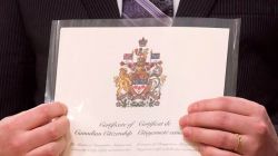 Person in a dark suit holds a Canadian Citizenship certificate inside a protective plastic sleeve, front view of the crest visible.