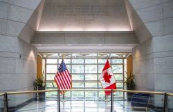 Interior lobby with American and Canadian flags standing near large windows, marble walls, and potted plants behind a glass railing.
