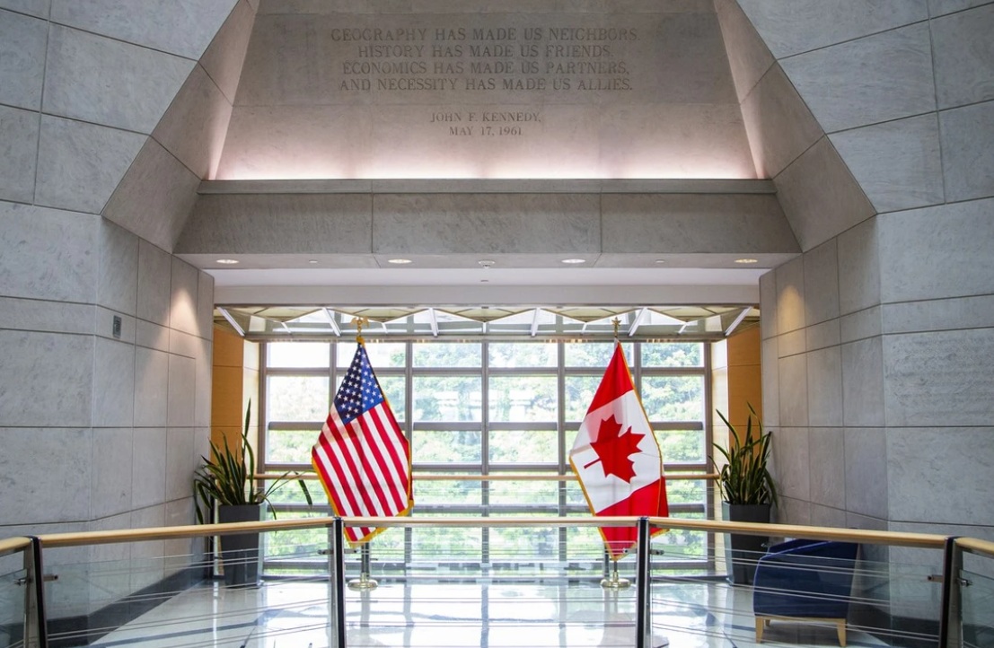 сша канада посолство | Наш Ванкувер Interior lobby with American and Canadian flags standing near large windows, marble walls, and potted plants behind a glass railing.