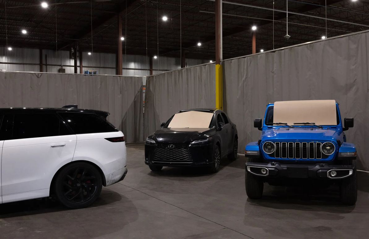 Three parked cars inside a warehouse-like garage: white SUV on the left, black sedan in the middle, and a blue Jeep Wrangler on the right with beige windshield covers.