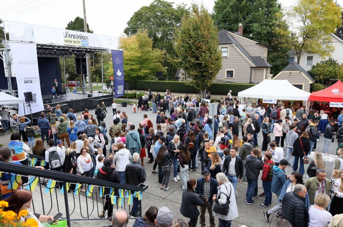 Crowd gathers at an outdoor festival with a stage labeled Ukrainian New West Fest and vendor tents nearby.