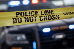 Police line tape reading 'Do Not Cross' stretched in front of flashing emergency vehicles behind.