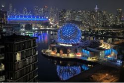 Nighttime city skyline with a glowing blue geodesic sphere on a waterfront promenade.