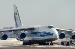 Large white cargo plane with blue stripe parked on a snowy airport tarmac; Russian flag on the tail and ground support equipment nearby.