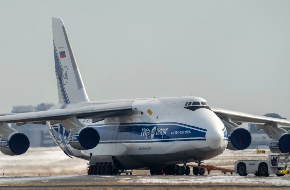 Large white cargo plane with blue stripe parked on a snowy airport tarmac; Russian flag on the tail and ground support equipment nearby.