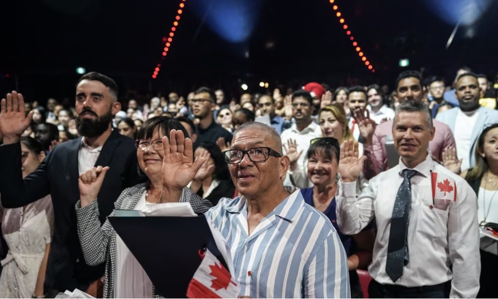 временные резиденты канада | Наш Ванкувер Crowd at an indoor event waving with hands raised; man in striped shirt in foreground holds a binder with a small Canadian flag.
