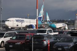 WestJet airplanes parked on the tarmac behind a chain-link fence, with a crowded parking lot in the foreground.