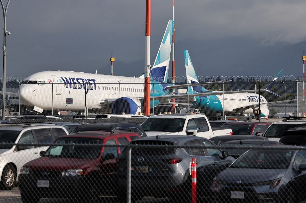 WestJet airplanes parked on the tarmac behind a chain-link fence, with a crowded parking lot in the foreground.