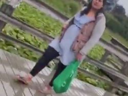 Woman standing on a wooden boardwalk outdoors, wearing a light blue dress and beige coat, carrying a bright green bag.
