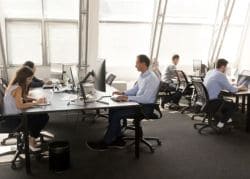Open-plan office with multiple workers at desks working on computers and laptops in a bright space.