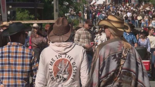 Two people in cowboy hats walk through a crowded outdoor event; one has a jacket that says 'Meet Me in the Desert.'