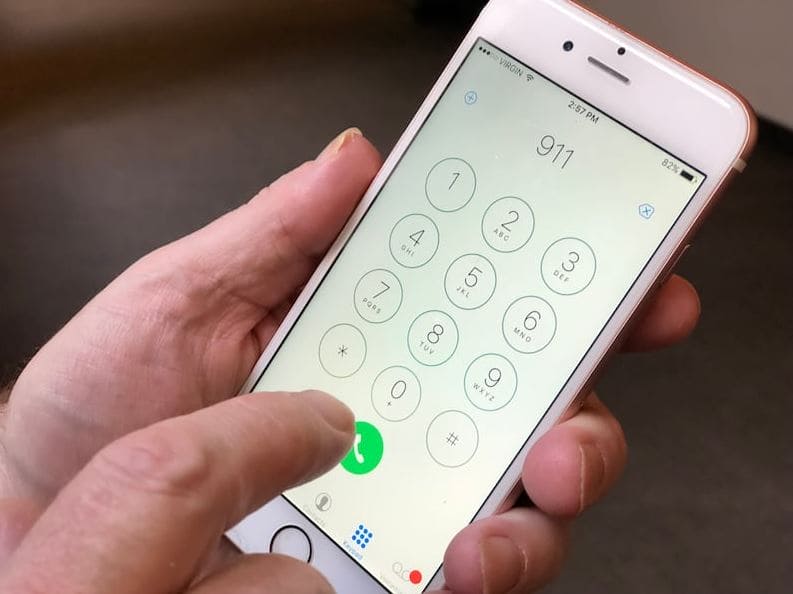 911 | Наш Ванкувер Hand holding a white iPhone showing a circular dial pad with a green call button at the bottom left, ready to dial a number