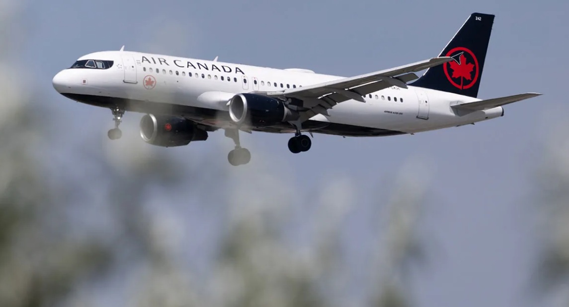 Air Canada passenger jet with gear down during approach against a blue sky with blurred foreground trees.
