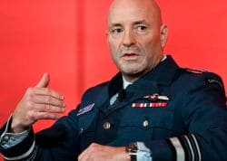 Male military officer in dress uniform speaking, with medals, against a bright red background.