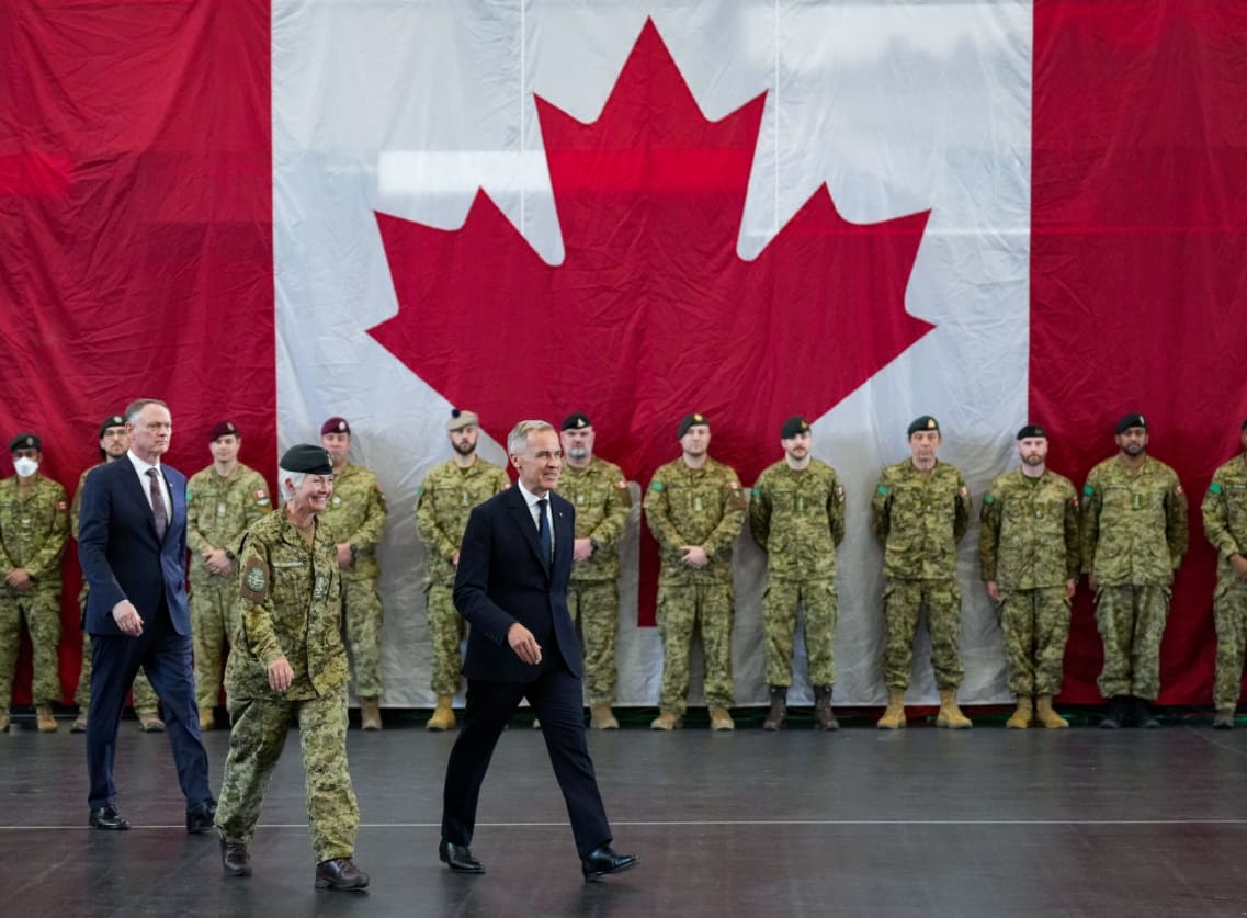 Dignitaries walk past soldiers in desert camouflage uniforms with a large Canadian flag backdrop behind them.