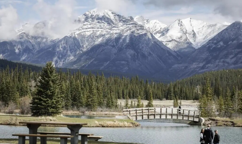 Snow-covered mountains rise behind a dense pine forest, with a wooden footbridge crossing a calm pond and a small group of people nearby.