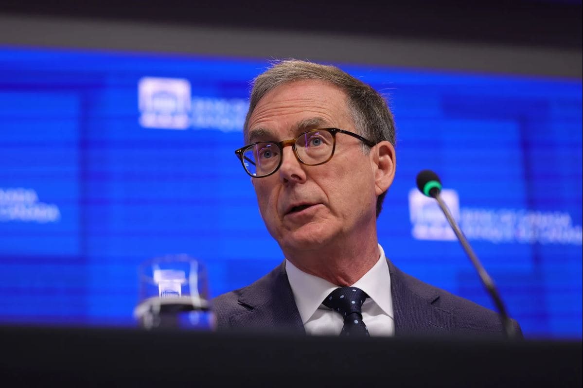 Банк Канады | Наш Ванкувер Man in suit and glasses at a podium during a press conference with a blue backdrop and a microphone nearby, speaking to an audience.