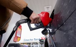 Person in a brown jacket refueling a black car with a red gas pump nozzle at a gas station.