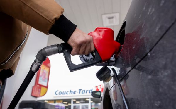 Person in a brown jacket refueling a black car with a red gas pump nozzle at a gas station.