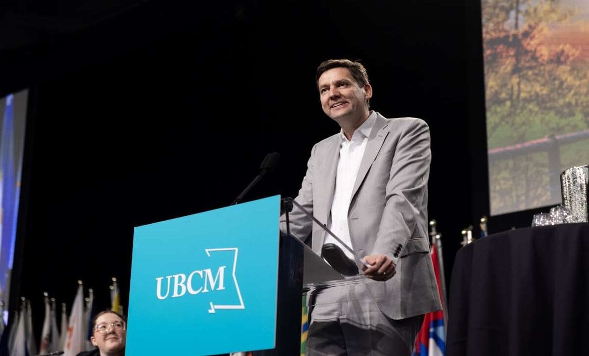 Британская Колумбия | Наш Ванкувер Man in a light gray suit speaks at a blue podium labeled 'UBCM' during a formal event, microphone in hand, stage with awards and flags nearby.