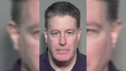 Mugshot of a middle-aged man with short dark hair, wearing a dark shirt, facing the camera against a gray backdrop