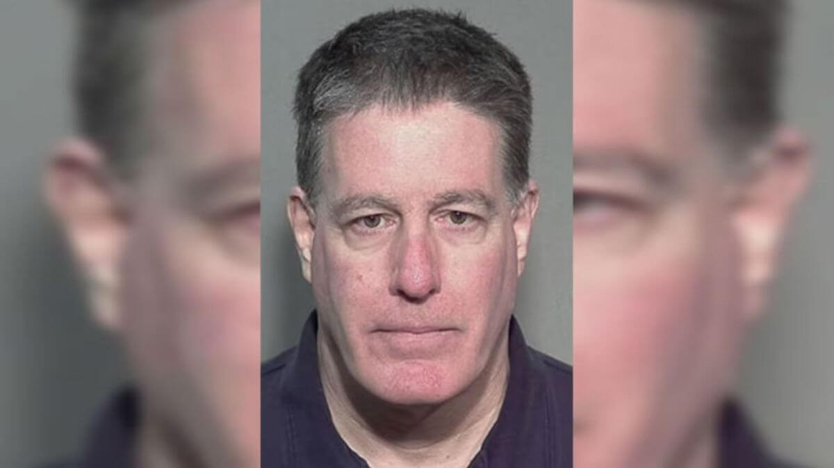 Mugshot of a middle-aged man with short dark hair, wearing a dark shirt, facing the camera against a gray backdrop