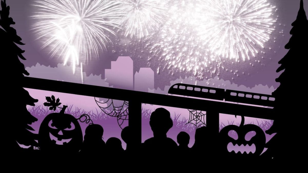 Silhouetted children and Halloween pumpkins on a balcony as fireworks light a purple night sky over a city skyline and train track.