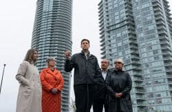 Five adults stand outdoors in front of two tall modern buildings; a man at a microphone speaks while others listen nearby.