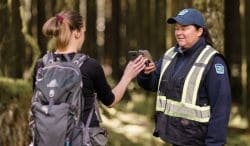 Hiker with a backpack hands a phone to a park ranger in a reflective-vest jacket in a forest path.