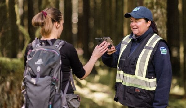 Hiker with a backpack hands a phone to a park ranger in a reflective-vest jacket in a forest path.