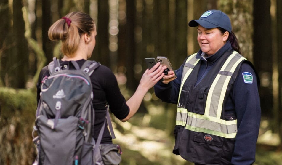 дневные пропуска | Наш Ванкувер Hiker with a backpack hands a phone to a park ranger in a reflective-vest jacket in a forest path.
