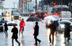 Rainy city street with pedestrians carrying umbrellas and a cyclist in a red rain suit crossing a flooded intersection; traffic behind them.