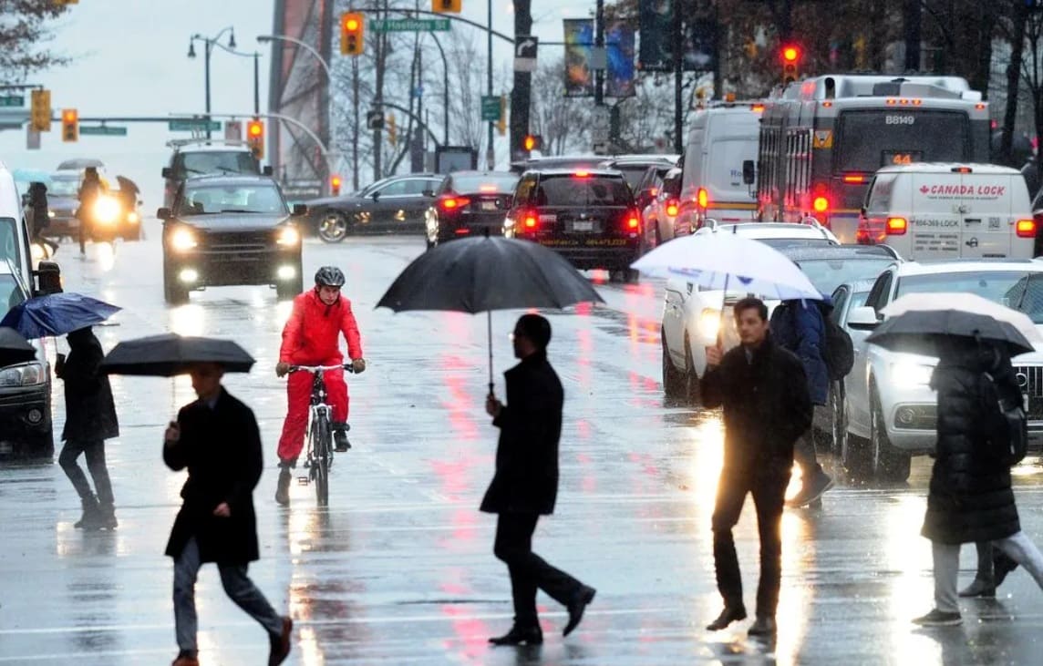 Rainy city street with pedestrians carrying umbrellas and a cyclist in a red rain suit crossing a flooded intersection; traffic behind them.