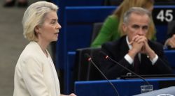 Blonde woman in a light blazer speaks at a podium with two microphones in a blue parliamentary chamber; a man in a suit sits behind with his hands clasped.