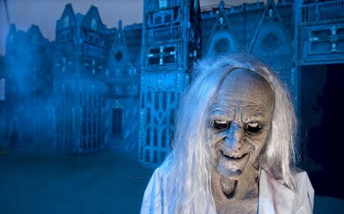 Terrifying witch prop with pale gray skin, long white hair, and a menacing grin in a blue-lit haunted setting behind her.