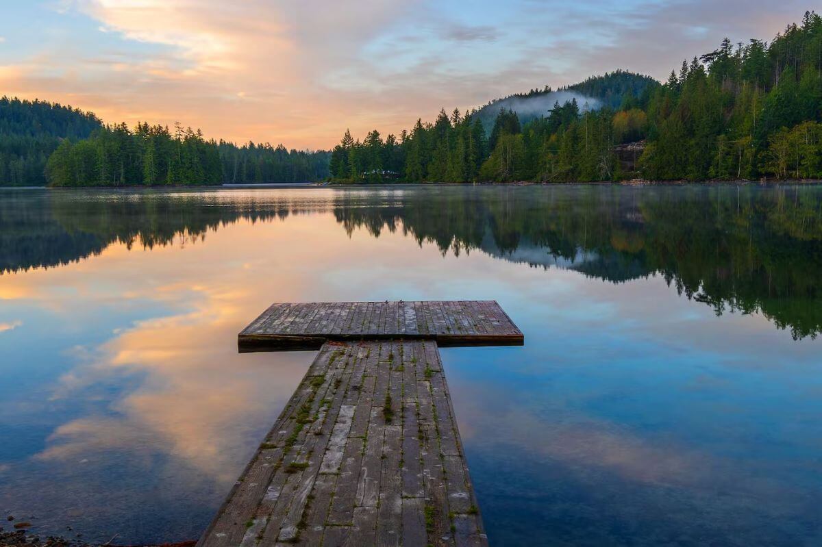 Wooden dock extending into a calm lake at sunset, with forested hills and a colorful sky reflected in the water.