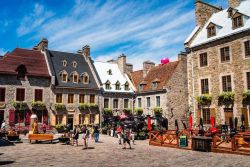 Quaint cobblestone square with stone buildings, outdoor cafe seating, and people strolling under a bright blue sky.