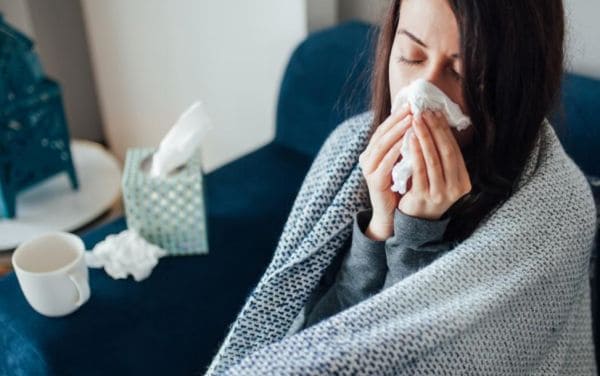 Woman wrapped in a blanket on a couch, sneezing into a tissue held to her nose. A tissue box and mug are on the table beside her.